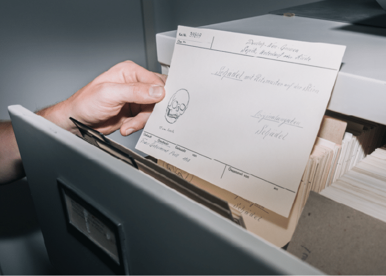 Hand legt eine beschriftete Karte in eine Archivschachtel mit Papieren